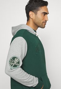 Sudadera verde y gris con un bolsillo canguro, bordada con "New York" en el frente y un logo de los Yankees en la manga izquierda. Capucha con cordón.