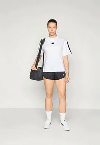 Ženska stoji oblečena v belo Adidas majico, črne kratke hlače, bele Adidas nogavice in čevlje, ter nosi črno športno torbo na belem ozadju.