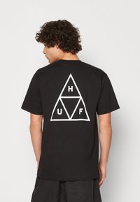 Czarny bawełniany t-shirt z białym geometrycznym wzorem trójkąta z literami H, U i F w środku. Krótkie rękawy i luźny krój.