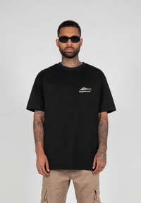 Sort bomulds t-shirt med korte ærmer og rund hals. Har et lille hvidt og guld logo på venstre bryst. Modellen bærer mørke solbriller.