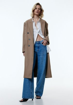 Lang beige jas met zwarte knopen, witte blouse en wijde blauwe jeans. Zwarte open-toed haksandalen. Egaal witte achtergrond.