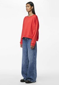 Sudadera roja de gran tamaño con mangas anchas, combinada con pantalones de mezclilla de pierna ancha en azul claro. Textura suave, corte casual y diseño minimalista.