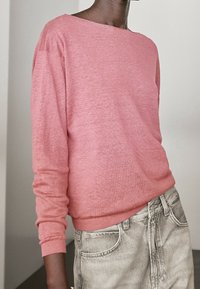 Różowy sweter z długim rękawem i dekoltem typu łódka, z prążkowanymi mankietami i dołem, noszony na jasnoszarych dżinsach, prezentujący miękką, teksturowaną tkaninę.