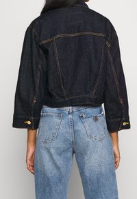 Tmavě modrá cropped džínová bunda s špičatým límcem, dlouhými rukávy a detaily se žlutým prošitím. Doplněno světle modrými džíny s vysokým pasem.