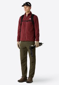 Giacca in pile bordeaux, pantaloni cargo oliva, scarponi da trekking marroni. Gli accessori includono guanti e un cappellino nero. Persona che tiene dei bastoncini da trekking.