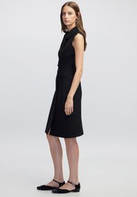 Schwarzes ärmelloses Kleid mit Kragen, vorderen Knöpfen und Rock bis zum Knie. Glattes Material mit seitlichem Schlitz.