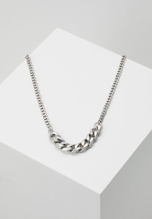 Collier en chaîne argentée avec des maillons alternant entre épais et fins, présenté sur un piédestal géométrique blanc.