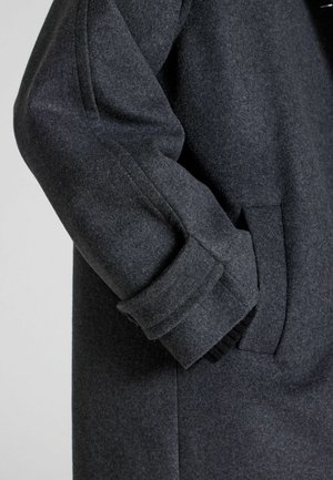 Tummanharmaa villakangastakki, jossa on istuva siluetti, näkyvät hihansuut ja sivutasku. Pehmeä rakenne ja voimakkaasti muotoiltu design.