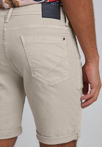 Pantaloncini beige in cotone con orli risvoltati, dotati di tasca applicata e una targhetta in pelle nera sulla cintura. Tessuto liscio e vestibilità sartoriale.