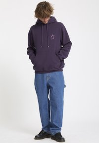 Purple hoodie with a star graphic on the chest, featuring a drawstring hood and front pocket. Paired with loose blue denim jeans and black shoes.