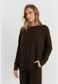 Maglione marrone lavorato a maglia con colletto rotondo, maniche lunghe e polsini a coste. Il tessuto appare morbido con una vestibilità comoda e tasche laterali.