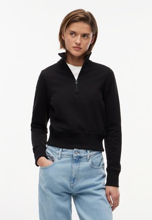 Ung kvinde med kort hår iført en sort hættesweater med lynlås over en hvid top og lyseblå jeans, stående med den ene hånd i lommen.
