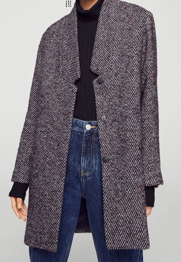 Femme portant un long manteau texturé gris et blanc sur un col roulé noir et un jean bleu taille haute, debout devant un fond uni.