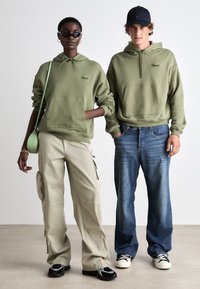 Dos personas visten sudaderas de color verde oliva a juego con un bolsillo frontal, junto con pantalones cargo beige y jeans azules, todo esto sobre un fondo neutro.