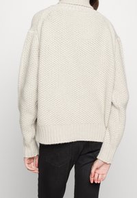 Jasnoszary dzianinowy sweter z wysokim kołnierzem, grubą fakturą, opuszczonymi ramionami oraz ściągaczami na mankietach i dole, noszony na czarnych dżinsach.