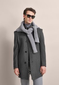 Grauer Wollmantel mit tailliertem Schnitt, schwarzen Knöpfen und Kapuze. Kombiniert mit einem hellgrauen ribbelnden Schal und schwarzen Sonnenbrillen.