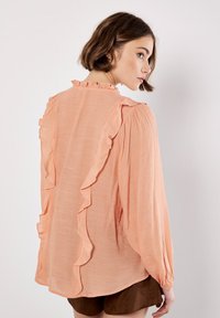 Blusa de color melocotón con acentos de volantes en los hombros y el escote. Mangas largas con puños fruncidos y una tela ligera y texturizada.