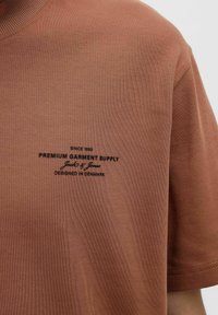 Trumpas rankovėmis, rūdžių spalvos marškinėliai, pagaminti iš minkštos medvilnės, su juodu logotipu krūtinės srityje ir su reljefiniu apykaklė.