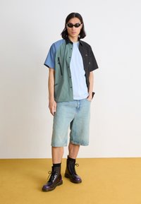 Short-sleeve, multi-colored shirt with zip pockets, paired with light blue t-shirt and denim shorts. Worn with glossy black and purple boots.