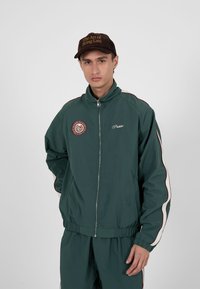 Jeune homme portant une veste zippée vert foncé et un pantalon assorti avec des bandes blanches et rouges, ainsi qu'une casquette brune avec l'inscription « The Art of Being Late ».