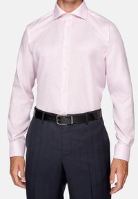 Man som bär en ljusrosa skjorta instuckna i mörka randiga byxor med svart läderbälte, stående mot vit bakgrund.