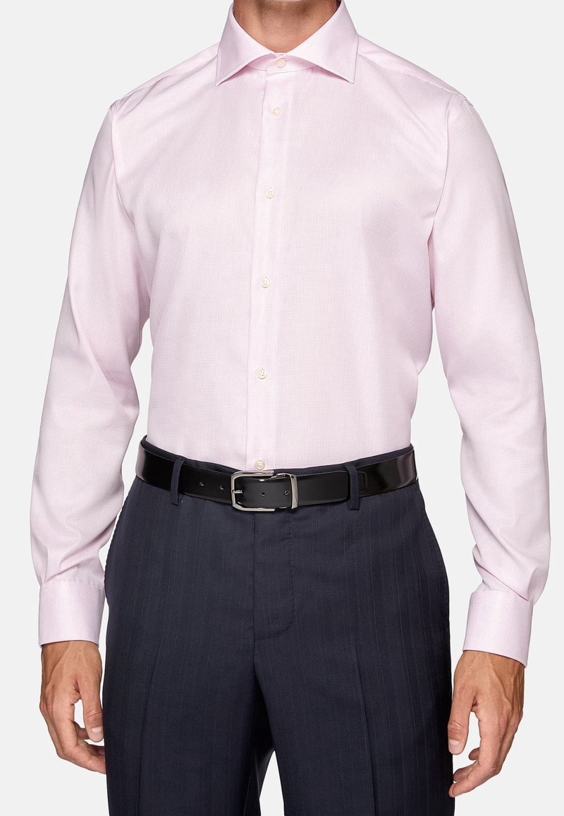 Man som bär en ljusrosa skjorta instuckna i mörka randiga byxor med svart läderbälte, stående mot vit bakgrund.