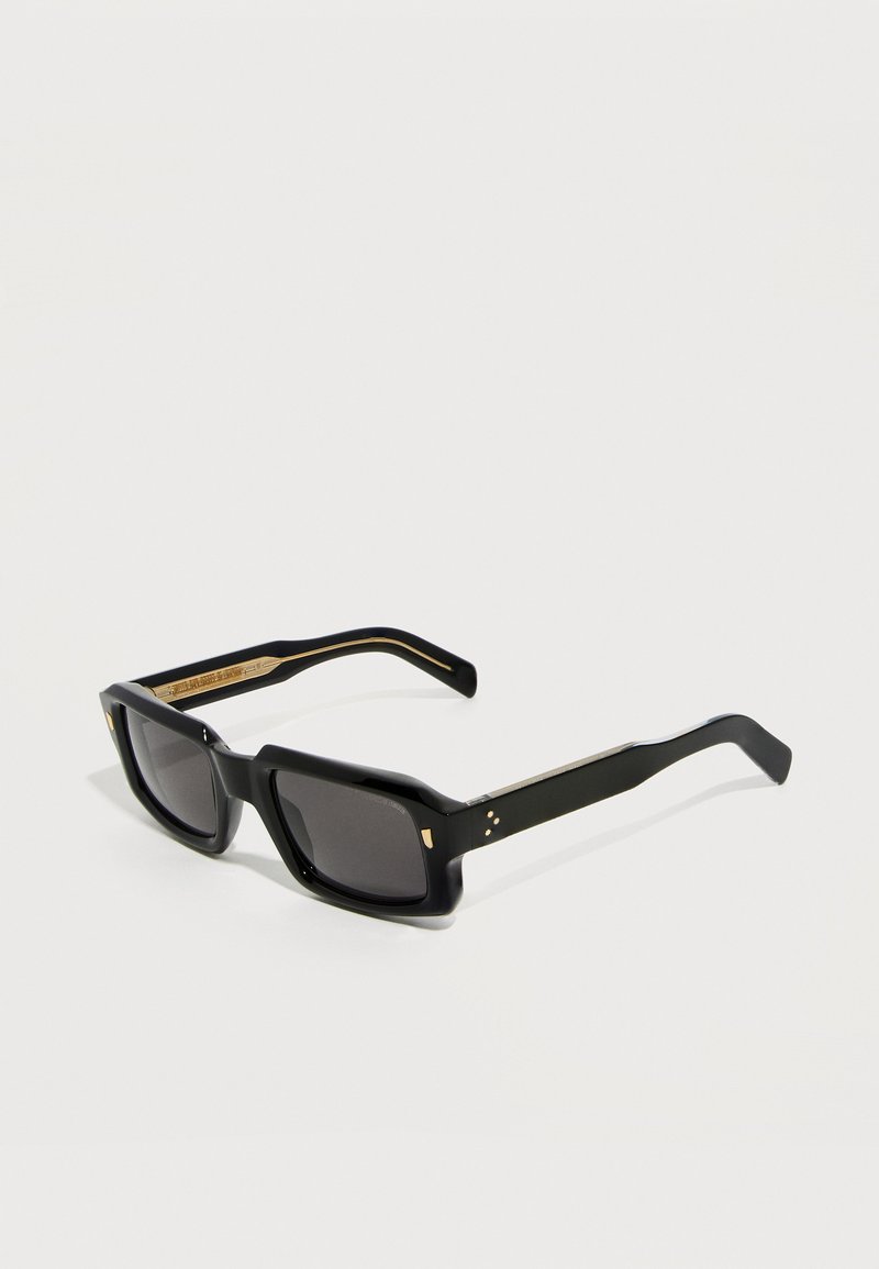 Μαύρα ορθογώνια γυαλιά ηλίου με χοντρές κορνίζες και σκούρους φακούς, με μικρές χρυσές λεπτομέρειες στις γωνίες και τα πλαϊνά τους.