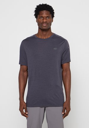 Homme aux cheveux courts et bouclés portant un t-shirt uni gris foncé à manches courtes et un pantalon gris clair sur fond blanc.
