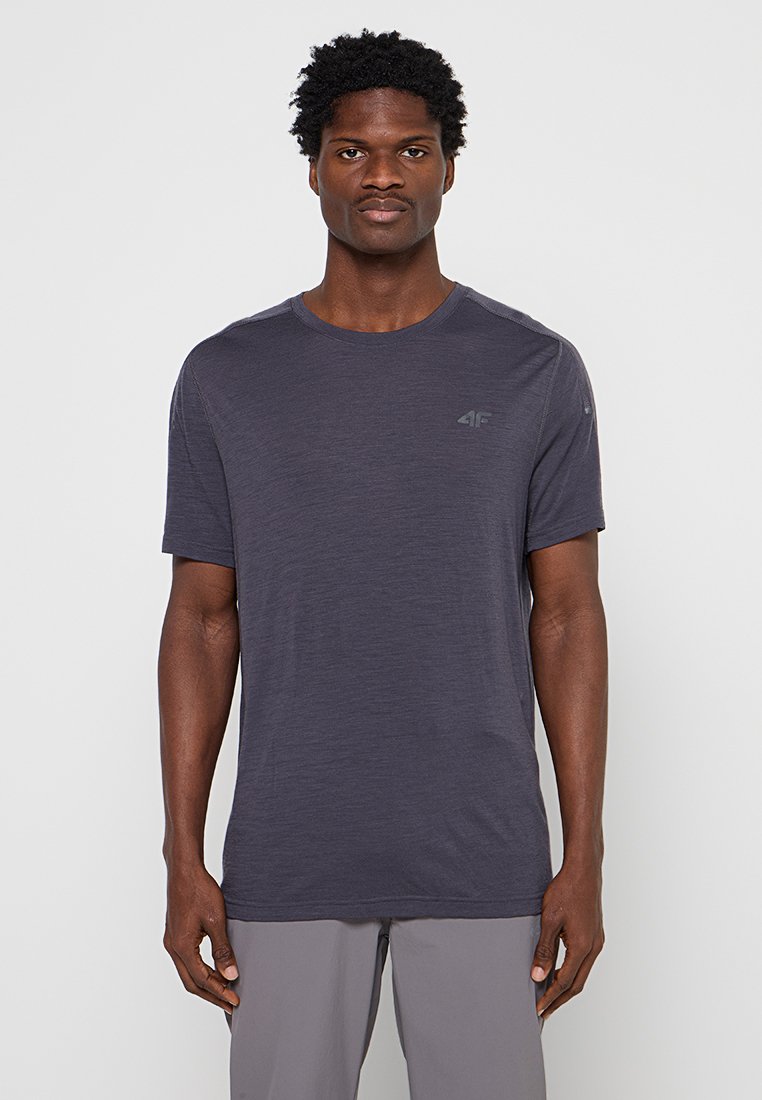 Homme aux cheveux courts et bouclés portant un t-shirt uni gris foncé à manches courtes et un pantalon gris clair sur fond blanc.