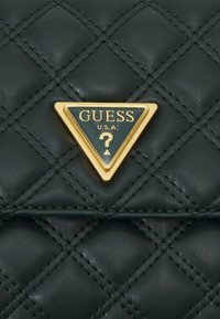 Sac en cuir matelassé vert avec une plaque logo triangulaire dorée portant le texte "GUESS U.S.A." et un motif point d'interrogation.