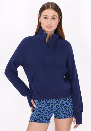 Mørkeblå zip-up sweatshirt med høj krave og kengurupoket, kombineret med shorts i blå leopardmønster. Blødt stof, afslappet pasform.