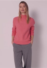 Růžový pletený svetr s vysokým límcem a žebrovaným lemem, v kombinaci se šedými pruhovanými kalhotami. Měkký materiál, volný střih a mírně nafouklé rukávy.