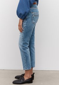 Jeans de mezclilla de color azul claro con corte recto, detalles desgastados en las rodillas y largo recortado. Combinados con mocasines negros, mostrando un look casual.