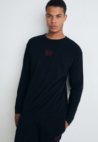 Cămașă neagră cu mâneci lungi, având un logo dreptunghiular roșu cu textul "HUGO" pe piept, confecționată dintr-un material fin, asortată cu pantaloni negri.