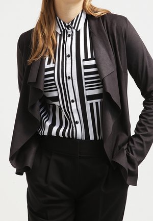 Persoon draagt een zwarte gedrapeerde open blazer over een zwart-wit verticaal gestreept overhemd met knopen, ingestopt in een zwarte broek.