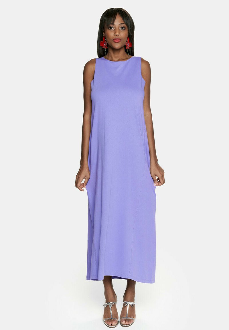 Długa sukienka bez rękawów w lawendowym materiale o luźnym kroju. Posiada wysoki dekolt i boczne kieszenie; noszona z srebrnymi obcasami.