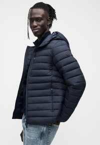 Μπουφάν ναυτικού μπλε με αποσπώμενη κουκούλα, καπιτονέ σχέδιο, τσέπες με φερμουάρ και στρογγυλό λογότυπο στο αριστερό μπράτσο. Υφή υφάσματος με τελείωμα.