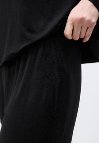 Gros plan sur une main soulevant une chemise noire pour révéler un pantalon noir avec un détail en dentelle florale verticale sur le côté.