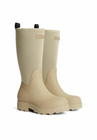 Beige kumisaappaat, jotka ulottuvat polveen, rakenne on teksturoitu, pinta on sileä ja ylhäällä on mustat vetonauhat. Minimalistinen muotoilu, jossa on kohotuslogo.
