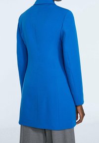 Manteau bleu vif sur mesure avec un col cranté, des manches longues et une silhouette ajustée, fabriqué en tissu lisse, avec des coutures nettes et minimalistes.