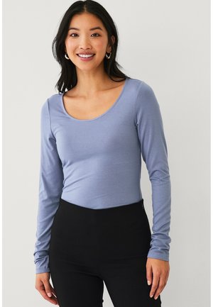 Smilende kvinde med langt mørkt hår iført store øreringe, en lyseblå langærmet bluse med rund hals og sorte bukser med høj talje.