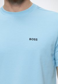 T-shirt bleu clair à col rond avec le logo texte noir "BOSS" sur la poitrine gauche, porté par une personne visible du cou jusqu'au haut du torse.