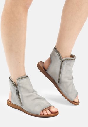 Füße in hellgrauen Sandalen mit offenem Zehenbereich, seitlichen Reißverschlüssen und flachen braunen Sohlen vor weißem Hintergrund.