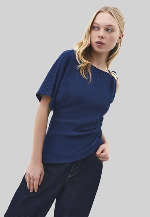 Jonge vrouw die een gestructureerde marineblauwe top draagt met één mouw en gouden schouderdetail, gecombineerd met donkere jeans met wijd uitlopende pijpen.