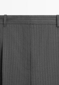 Pantalon gris à fines rayures avec passants pour ceinture et fermeture avant dissimulée, montrant des coutures verticales détaillées et la texture du tissu.