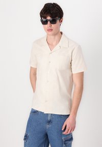 Kurzarm, hellbeige Hemd mit Knöpfen und einem dezenten Muster sowie einer einzelnen Brusttasche, kombiniert mit blauen Denim-Cargoshorts.