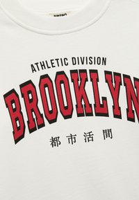 Biały sweter z okrągłym dekoltem z wyraźnym czarnym i czerwonym napisem "BROOKLYN" oraz "ATHLETIC DIVISION" powyżej w łuku.
