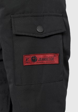 Black cargo pants with a side pocket featuring a black button and a red label that reads "JEANSCODE" with additional printed details.