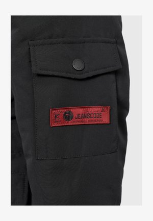 Schwarze Cargo-Hosen mit einer seitlichen Tasche, die einen schwarzen Knopf und ein rotes Etikett mit der Aufschrift "JEANSCODE" sowie zusätzlichen bedruckten Details aufweist.