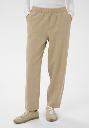 Personne portant un pantalon beige ample avec une taille élastique et des baskets beige, les mains dans les poches, debout sur un fond blanc.